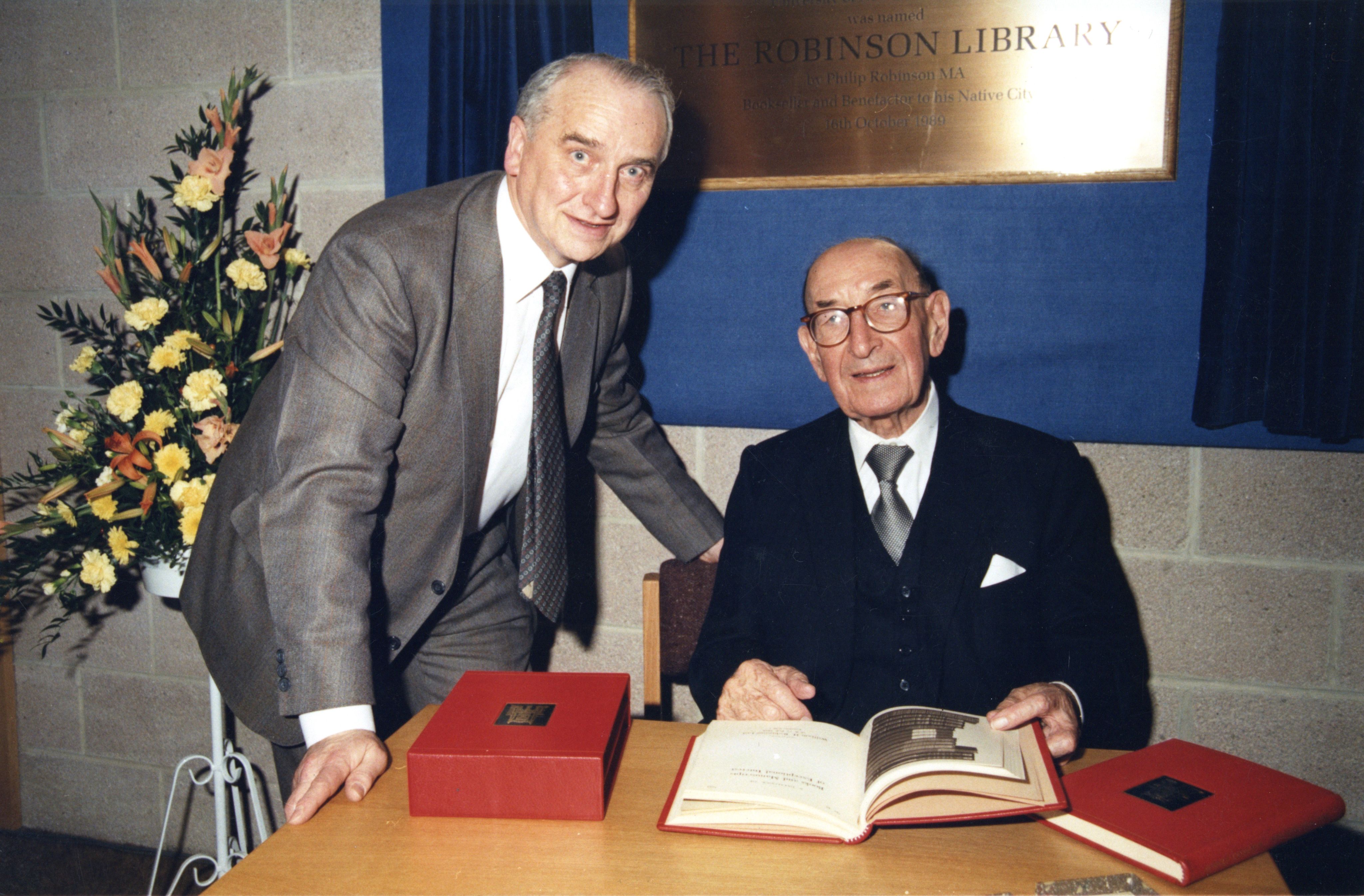 Two men seated in front of a brass plague with books on a table.